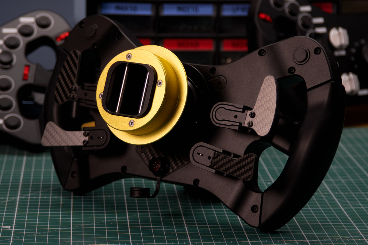 A close-up of a black racing steering wheel with a prominent yellow QR and textured grips, resting on a green cutting mat with another steering wheel blurred in the background.