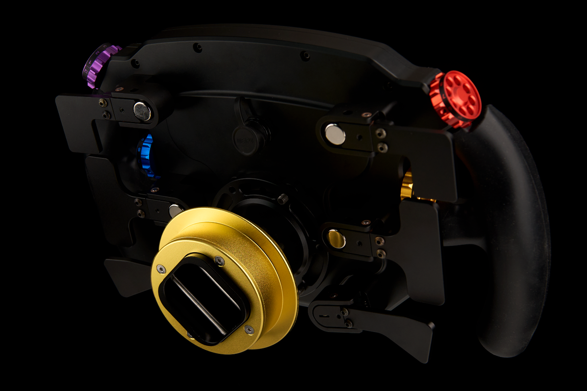 A high-performance racing wheel with a black frame, a prominent gold QRl, a black grip, and colored dials in purple, blue, and red, set against a solid black background.