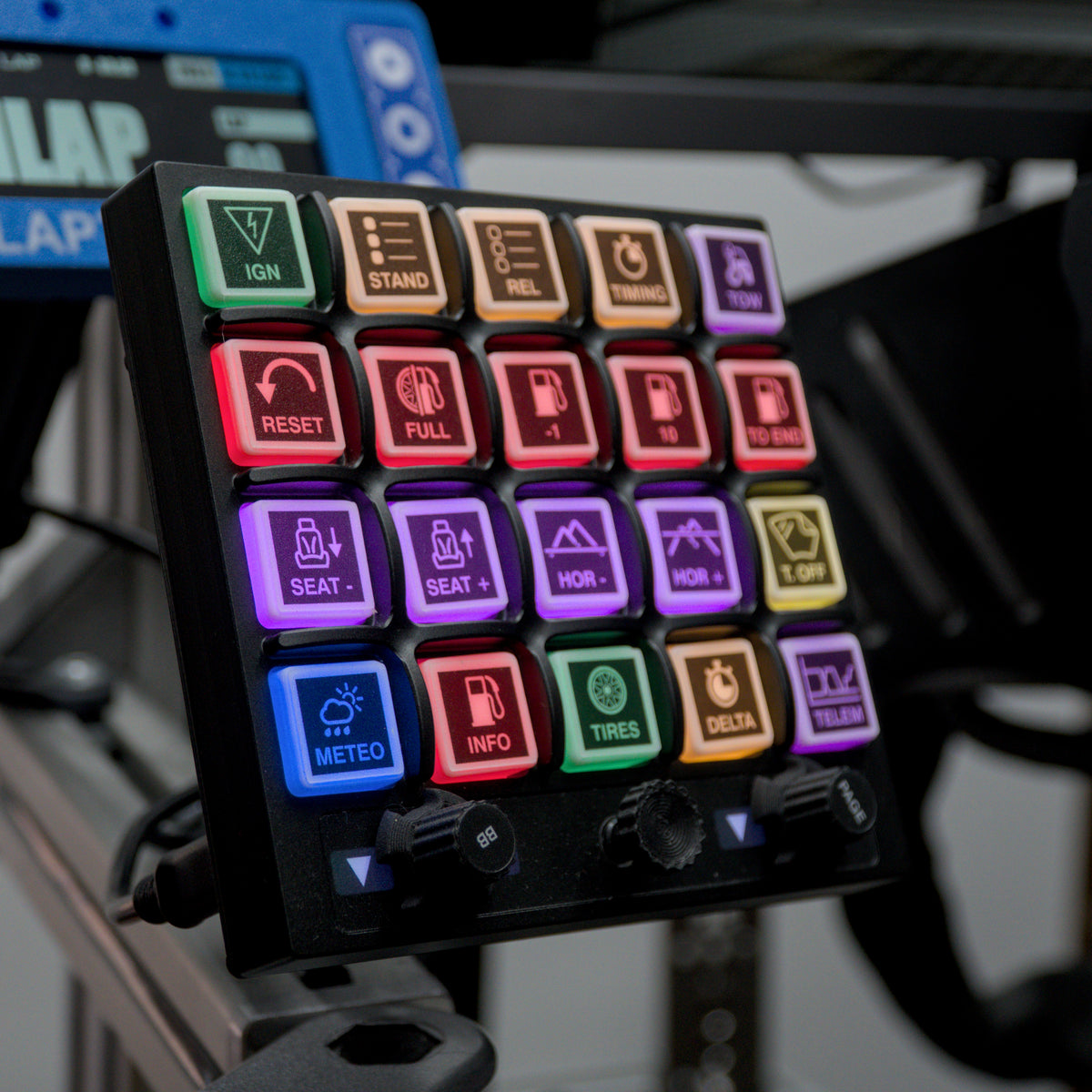 A close-up of a black electronic control panel with 24 colorful illuminated buttons arranged in a grid, each labeled with different functions such as IGN, STAND, BELL, CHARGE, SEAT-, METEO, and TIRES. The panel is mounted on a stand, with part of a blue device visible in the background.